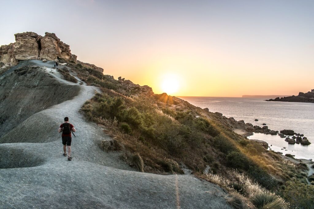 Randonneur sur un sentier côtier rocheux au coucher du soleil, mer en arrière-plan.