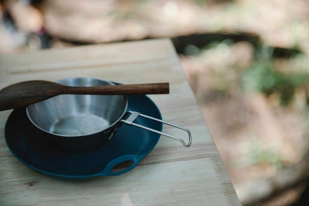 Ustensiles de camping: casserole en métal, cuillère en bois sur un plateau bleu.