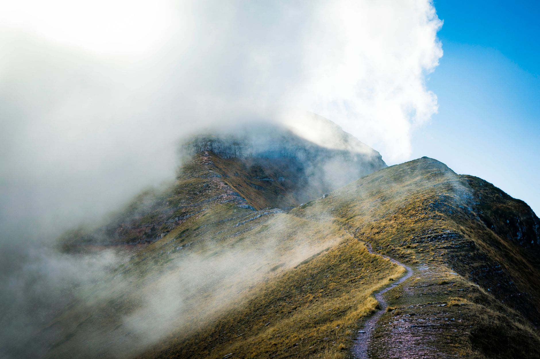 Sentier rocailleux serpentant sur une crête montagneuse, partiellement couverte de nuages.