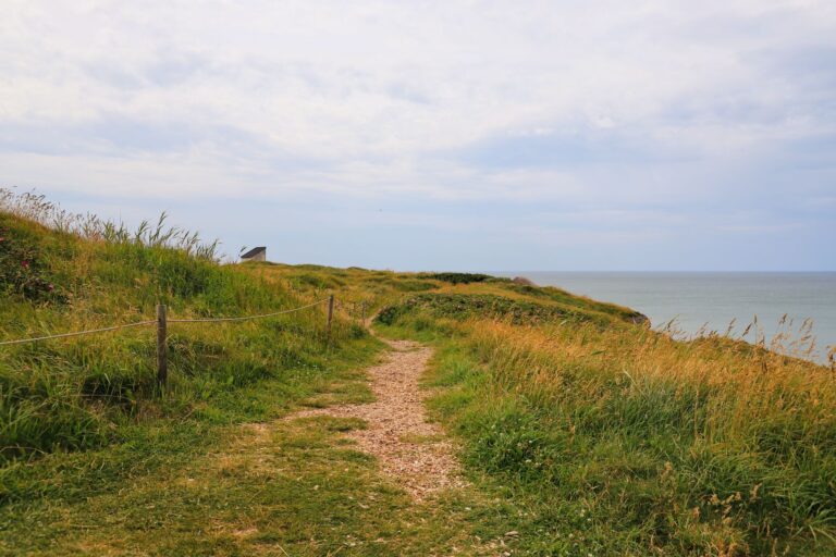Chemin de terre sur une colline herbeuse surplombant la mer, cabane lointaine.