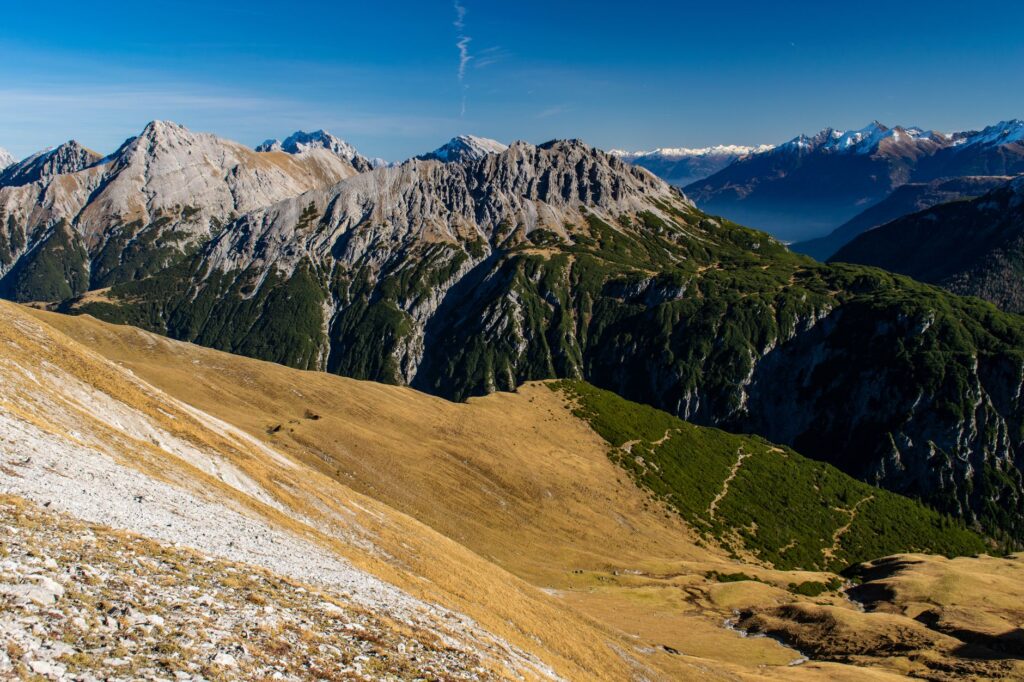 Massif montagneux rocheux et boisé, pentes herbeuses au premier plan sous ciel bleu.